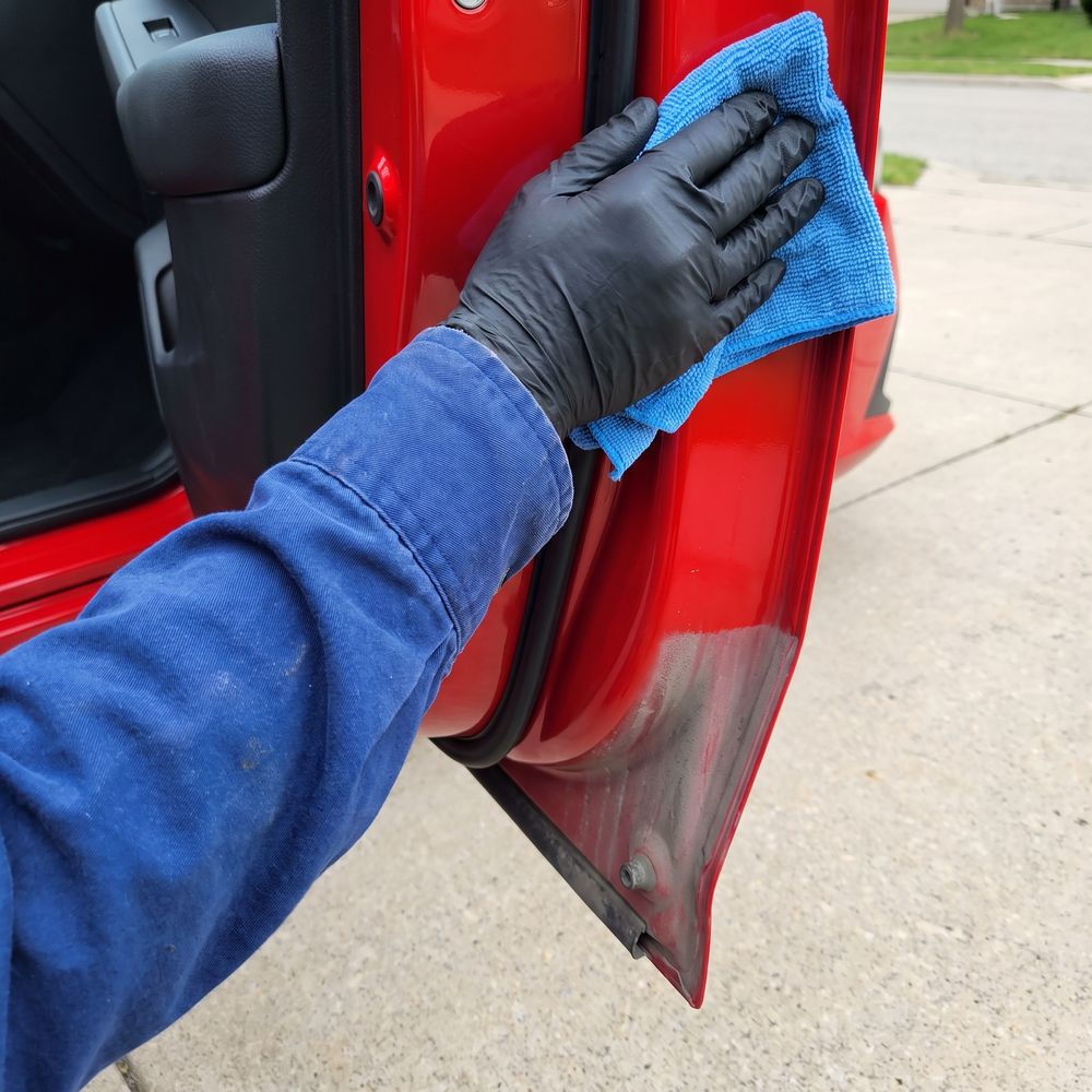 A gloved hand in a blue work sleeve wiping the door jamb and hinge of a red Honda Civic with a blue microfiber towel. The top of the jamb is clean and glossy, the bottom still gray with road grime.