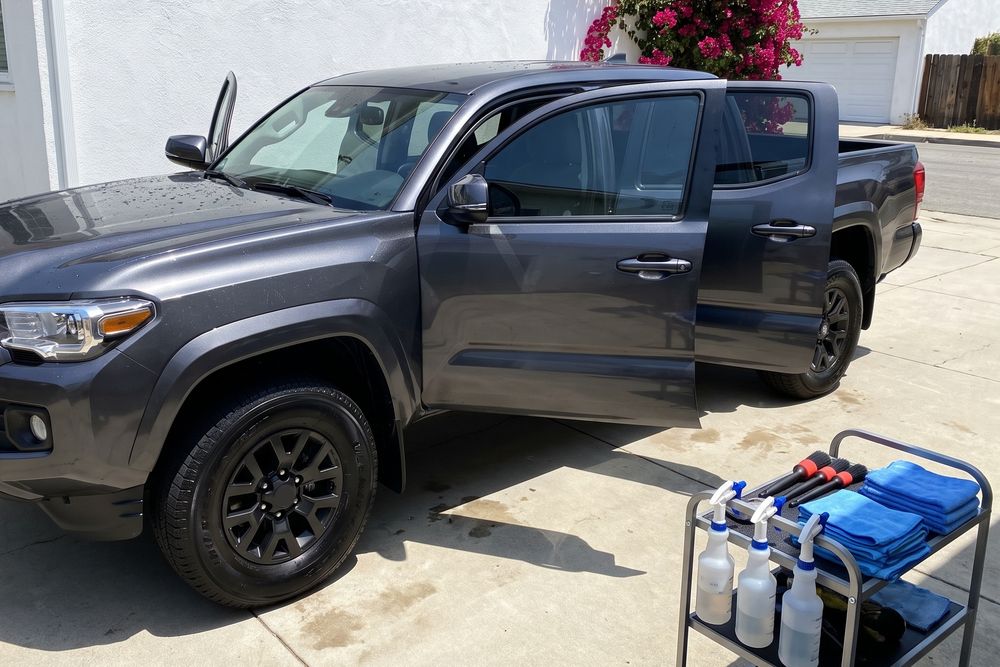 A dark gray Toyota Tacoma pickup on a driveway beside a white stucco house with all its doors open, paint freshly washed, tires dressed black, and a cart of spray bottles and blue microfiber towels next to it. Pink bougainvillea grows on the wall behind.