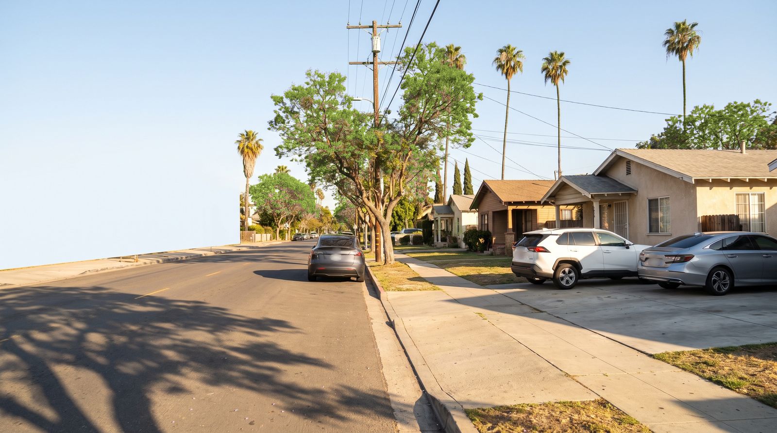 A Los Angeles residential street in late afternoon sun: bungalows with short driveways, a white SUV and a silver sedan parked in them, a gray car at the curb, green street trees, palm trees and power lines.